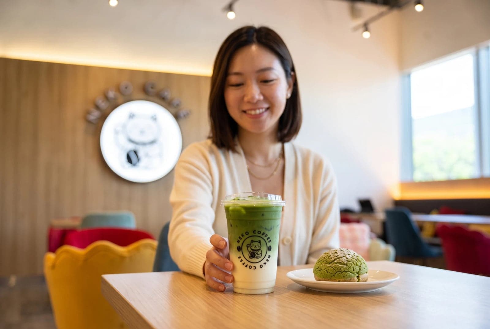 A smiling guest at Neko Cafe with an iced matcha latte in a Neko Coffee cup and a green matcha puff on a plate, the glowing lucky-cat sign on the wall behind